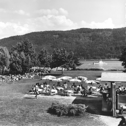 Seeufer mit Ausflugsboot und vielen Gästen auf Liegestühlen und an Tischen mit Sonnenschirmen